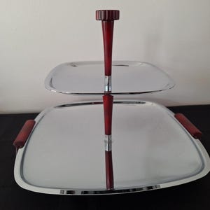 May include: A two-tiered serving tray with a polished chrome finish. The tray has a square shape with rounded corners and a red handle on top. The handles on the sides are also red. The tray is designed for serving food or displaying items.