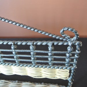 Pu&ograve; includere: Un porta riviste in metallo nero con un ripiano in vimini intrecciato. Il porta riviste ha un design decorativo, attorcigliato e una parte superiore curva.