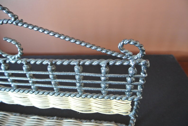 Pu&ograve; includere: Un porta riviste in metallo nero con un ripiano in vimini intrecciato. Il porta riviste ha un design decorativo, attorcigliato e una parte superiore curva.