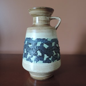 May include: A brown and white ceramic vase with a blue and green design. The vase has a handle and a wide mouth.