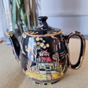 Könnte beinhalten: Schwarze Teekanne mit goldenen Verzierungen und einem farbenfrohen asiatisch inspirierten Design. Die Teekanne hat einen runden Korpus und einen Ausguss auf einer Seite. Der Griff befindet sich auf der gegenüberliegenden Seite.