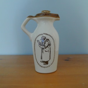 Puede incluir: Una jarra de cerámica blanca con un asa marrón y un diseño marrón de un hombre con un abrigo. La jarra tiene un tapón de corcho.
