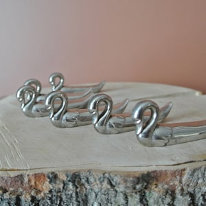 May include: Set of five silver swan napkin rings. The rings are shaped like swans with their necks curved and heads facing forward. The rings are arranged in a row on a wooden surface.