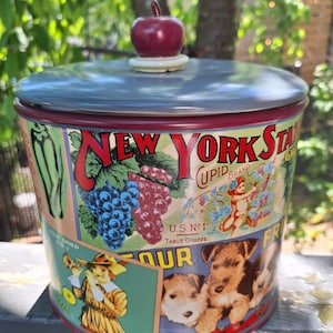 May include: A vintage-style ceramic cookie jar with a red apple lid. The jar features colourful illustrations of grapes, a cherub, and dogs, with the text "New York State Cupid Brand U.S. No. 1 Table Grapes Our Friend".