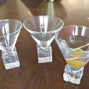 May include: Three clear glass martini glasses with square bases. Two glasses are empty and one glass contains a green olive and a red cocktail umbrella.