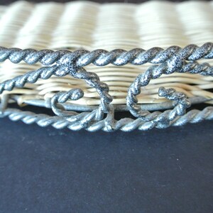 Pu&ograve; includere: Un cesto di vimini intrecciato con un bordo in metallo argentato. Il bordo presenta un design decorativo con volute e curve.