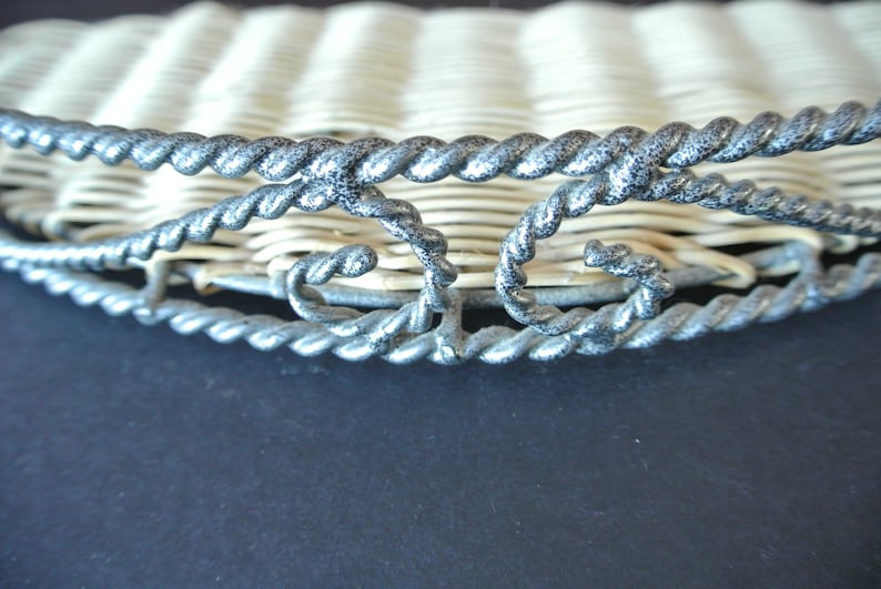 Pu&ograve; includere: Un cesto di vimini intrecciato con un bordo in metallo argentato. Il bordo presenta un design decorativo con volute e curve.