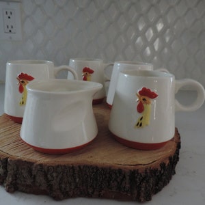 Op de afbeelding: Set van vijf witte keramische mokken met rode en gele haanontwerpen. De mokken hebben een rode rand en zijn gerangschikt op een houten schijf.