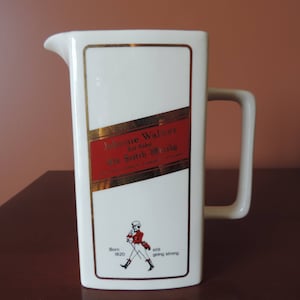 May include: White ceramic jug with a gold and red label that says "Johnnie Walker Old Scotch Whisky" and a small image of a man in a red jacket. The label also says "Born 1820 Still going strong".