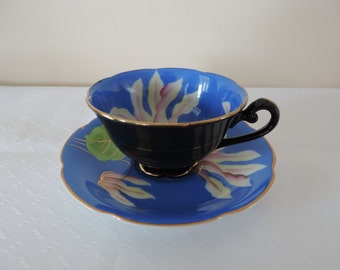 Puede incluir: Un juego de taza y platillo de té vintage. La taza es negra con interior azul y ribete dorado. El platillo es azul con un diseño floral. El juego es de porcelana y probablemente de mediados del siglo XX.