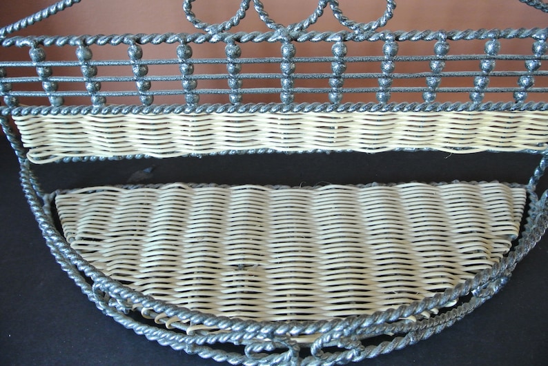 Pu&ograve; includere: Un ripiano a muro in metallo grigio con due ripiani in vimini intrecciato. Il ripiano ha un design decorativo a corda attorno ai bordi.