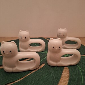 May include: Four white ceramic cat napkin rings. The rings are shaped like cats lying down with their heads turned to the side. The cats have black eyes and whiskers.