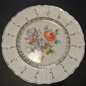 Puede incluir: Plato de cena de cerámica blanca con borde dorado y diseño floral con un iris morado, flores naranjas y amarillas. El plato tiene un borde elevado y texturizado.