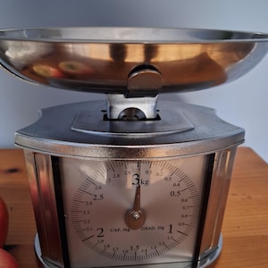 Peut inclure: Une balance de cuisine vintage en argent et chrome avec un cadran circulaire. La balance a un grand bol et une capacité de 3 kg. Le cadran affiche les mesures en kilogrammes et en grammes. La balance est sur une surface en bois.