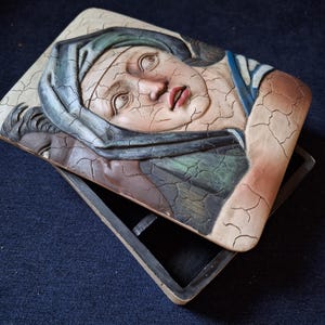 Op de afbeelding: Een houten doos met een scharnierend deksel. Het deksel heeft een reliëf, geschilderd beeld van een vrouwengezicht met een blauw-groene hoofddoek. De doos heeft een verweerde, antieke uitstraling.