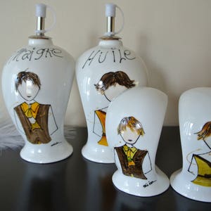 May include: Four white ceramic bottles with hand-painted designs of people. The bottles are labelled "Huile" and "Vinaigre".