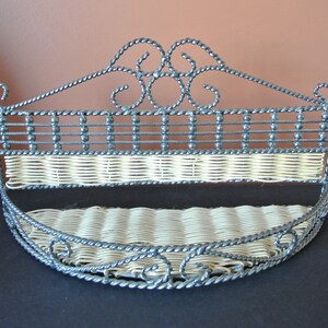 Pu&ograve; includere: Un ripiano a muro in metallo nero con un inserto in vimini intrecciato. Il ripiano ha un design decorativo a volute nella parte superiore e un fondo curvo.