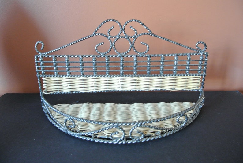 Pu&ograve; includere: Un ripiano a muro in metallo nero con un inserto in vimini intrecciato. Il ripiano ha un design decorativo a volute nella parte superiore e un fondo curvo.