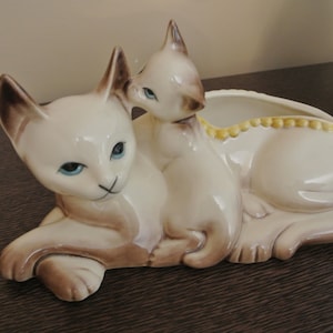 May include: A white ceramic planter shaped like two Siamese cats. The larger cat is lying down with its head turned to the side, while the smaller cat is curled up on its back. The planter has a yellow rim and a row of yellow dots around the edge.