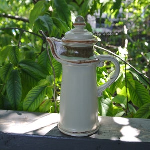 Pode incluir: Um jarro de cer&acirc;mica com tampa e al&ccedil;a. O jarro &eacute; branco com detalhes marrons e verdes. Est&aacute; sobre uma superf&iacute;cie de madeira.