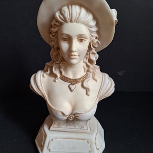 May include: A cream-coloured bust of a woman wearing a wide-brimmed hat. The sculpture features a detailed face, curly hair, and a necklace with a heart pendant. The bust is mounted on a decorative base.