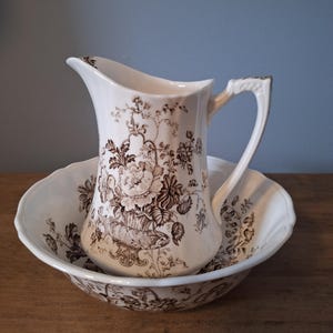 May include: A white porcelain pitcher and bowl set with a brown floral pattern. The pitcher has a handle and a spout. The bowl has a scalloped edge.