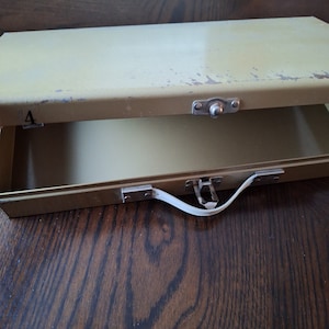 Könnte beinhalten: Eine vintage, hellgrüne Metall-Aufbewahrungsbox mit einem Griff und einem Verschluss. Die Box ist leicht abgenutzt und hat ein paar Kratzer auf der Oberfläche. Die Box ist offen und das Innere ist leer.