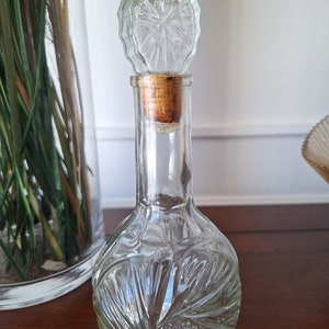 Puede incluir: Una botella de vidrio transparente con un tapón de corcho y una tapa de vidrio decorativa. La botella tiene un diseño de espiral y es perfecta para contener líquidos.