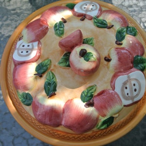 May include: A ceramic serving platter with an apple design. The platter has a light tan centre with red and white apples, green leaves, and a brown rim. The apples are arranged around the edge and centre of the platter.
