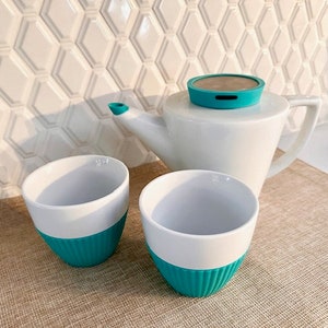 Puede incluir: Una tetera blanca con una tapa de color turquesa y dos tazas de té blancas con bandas de color turquesa. Las tazas de té y la tetera están sobre un mantel tejido marrón.
