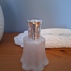 May include: A frosted glass oil burner with a silver lid featuring a floral design. The burner has a bell-shaped base.