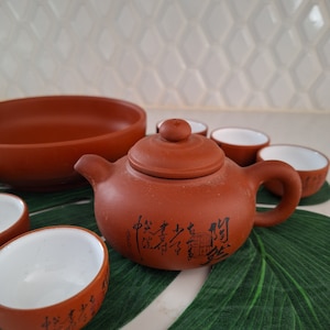 Puede incluir: Un juego de té de terracota con una tetera, un tazón grande y cuatro tazas pequeñas. La tetera tiene caracteres chinos en el lateral. El juego está sobre una alfombra verde en forma de hoja.