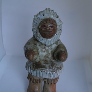 May include: A ceramic figurine of a person wearing a white hooded parka and brown boots. The person has a brown face and is smiling.