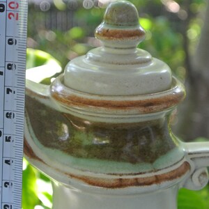 Pode incluir: Uma chaleira de cer&acirc;mica com um esmalte marrom, verde e branco. A chaleira tem uma tampa com um bot&atilde;o no topo e uma al&ccedil;a no lado.