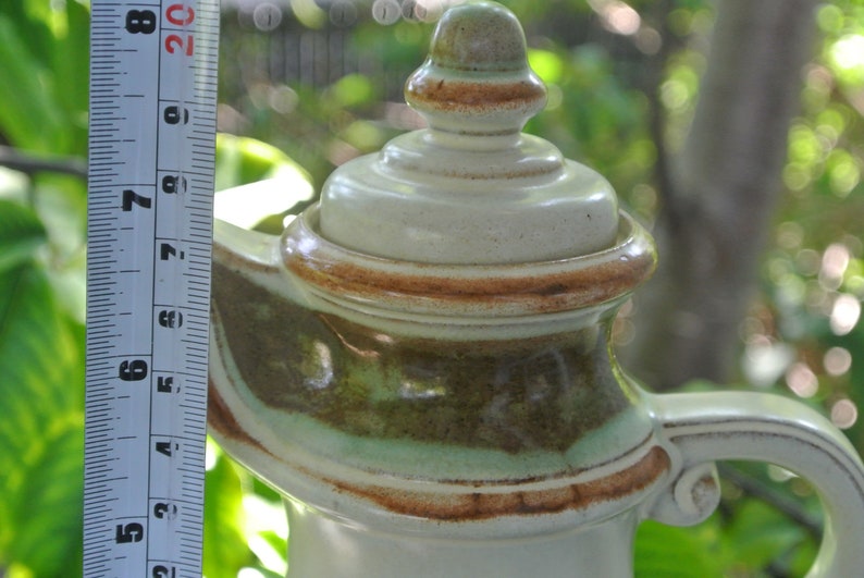 Pode incluir: Uma chaleira de cer&acirc;mica com um esmalte marrom, verde e branco. A chaleira tem uma tampa com um bot&atilde;o no topo e uma al&ccedil;a no lado.