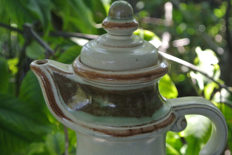 Pode incluir: Uma chaleira de cer&acirc;mica com um esmalte marrom, verde e branco. A chaleira tem uma tampa com uma pega no topo. A chaleira est&aacute; sentada sobre um fundo de folhas verdes.