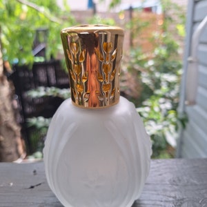 Puede incluir: Una botella de difusor de vidrio blanco esmerilado con una tapa de metal dorado que presenta un diseño floral decorativo. La botella tiene una forma redondeada y una superficie texturizada.