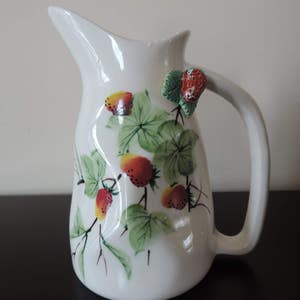 Puede incluir: Jarra de cerámica blanca con asa, decorada con un diseño pintado a mano de hojas verdes y fresas rojas. La jarra tiene una fresa 3D en el asa.
