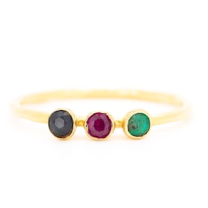 May include: A delicate gold ring featuring three round gemstones: a dark blue sapphire, a vibrant ruby, and a bright green emerald. The gemstones are set in individual bezels along the ring's band.