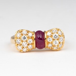 May include: A gold ring with a bow-shaped design. The bow's ends are encrusted with small, clear gemstones, while the center features a deep red, oval-shaped stone. The ring is set against a plain white background.