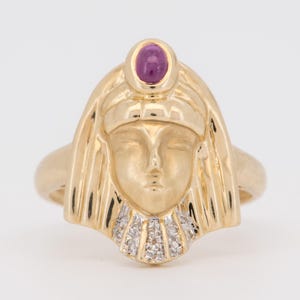 May include: A gold ring featuring a sculpted face with a smooth finish, topped with a single oval cabochon-cut purple gemstone at the forehead. The base of the face is adorned with a fan-shaped arrangement of small, clear, sparkling stones.