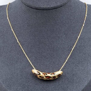 May include: A gold-toned necklace with a pendant shaped like a curved banana. The pendant has a leopard print pattern in brown and red. The necklace is displayed on a gray mannequin.