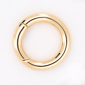 May include: A gold-colored, circular clasp with a hinged opening. The clasp is smooth and reflective, with a polished finish. The design is simple and modern, suitable for jewelry or accessories. The clasp is shown against a white background.