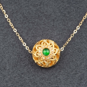 Könnte beinhalten: Eine goldfarbene Halskette mit rundem Anhänger. Der Anhänger hat ein durchbrochenes Design mit einem zentralen grünen Stein und umliegenden klaren Steinen. Die Kette ist ebenfalls goldfarben. Der Durchmesser des Anhängers beträgt ca. 2 cm.