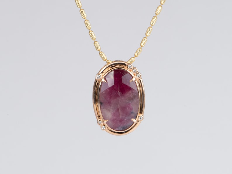 May include: A gold chain necklace with a pendant featuring a large, oval-shaped, pink and purple gemstone surrounded by smaller, clear gemstones.