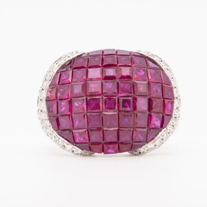 May include: An oval-shaped ring featuring a mosaic of square-cut rubies in a deep magenta hue. The ring is bordered by a band of small, sparkling diamonds set in a white metal, creating a striking contrast.