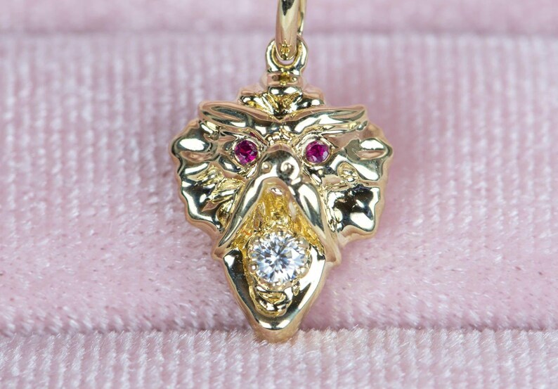 May include: Close-up of a gold dragon pendant featuring ruby eyes and a central diamond. The dragon's mouth is open, revealing the diamond. The pendant is intricately detailed and has a three-dimensional quality.  The gold is a bright, shiny yellow, and the rubies are a deep, rich red. The diamond is clear and sparkly. The pendant is shown against a pink velvet background.