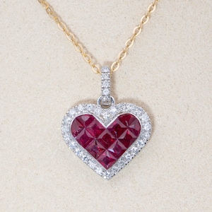 May include: A heart-shaped pendant necklace with a gold chain. The pendant features a central cluster of deep red gemstones, surrounded by a halo of smaller, clear gemstones. The pendant is approximately 2.5 cm in width.
