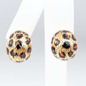 May include: A pair of gold-coloured oval earrings with a leopard print design. The earrings have black spots and orange accents on a gold background. The earrings are displayed on white earring stands.