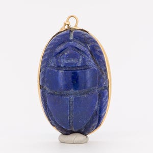 May include: A deep blue lapis lazuli scarab beetle pendant set in a gold-toned metal frame. The oval-shaped stone features carved details representing the head, wings, and body of a beetle, with a small gold-toned metal bail at the top for hanging.